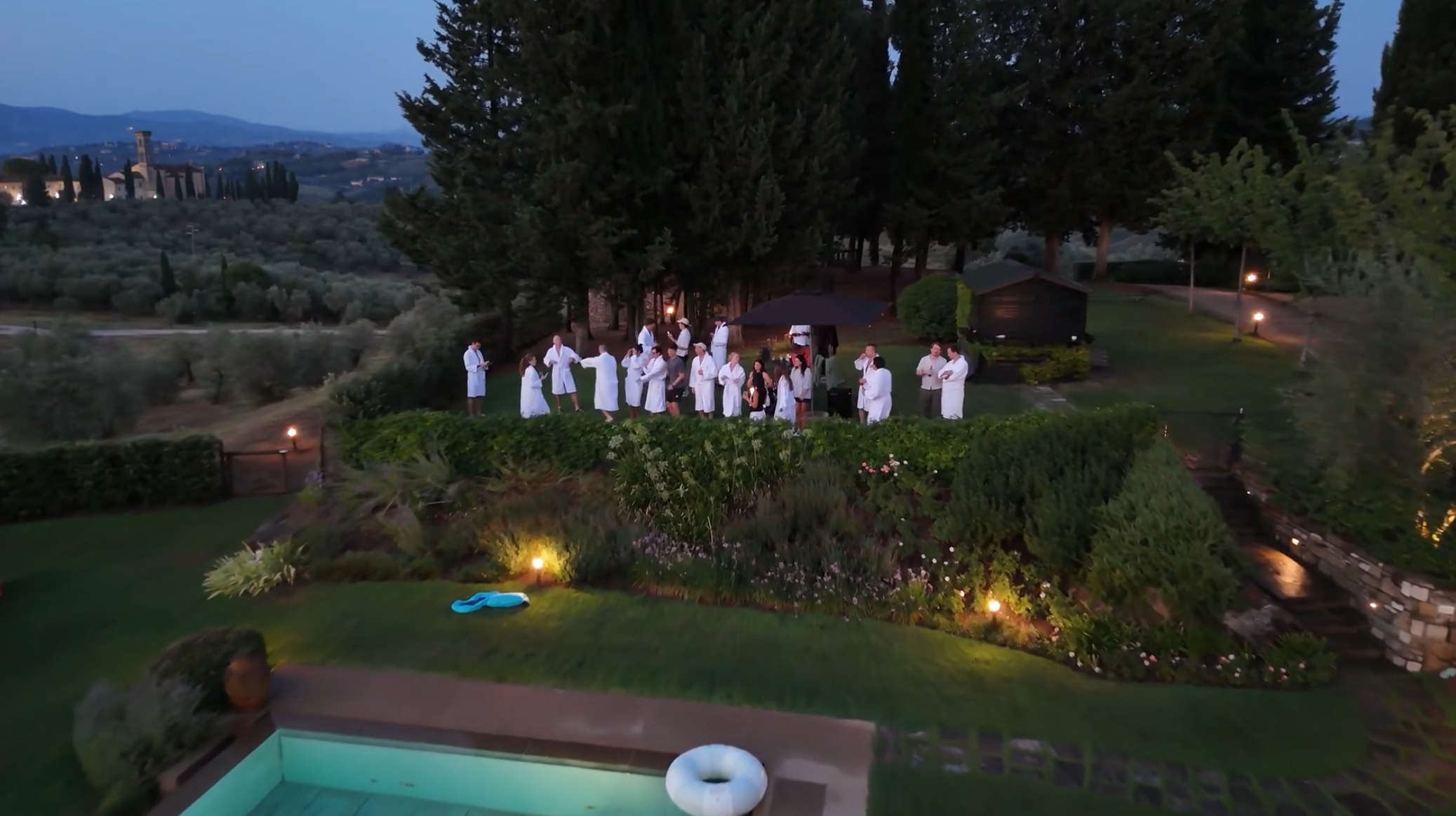Drone view of friends in bathrobes gathered in an Italian garden at dusk, with a pool in the foreground.