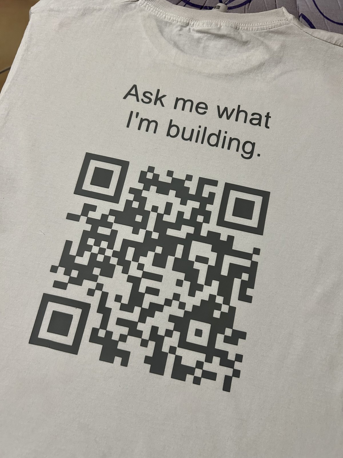 A white t-shirt printed with the words Ask me what I'm building and a large QR code.
